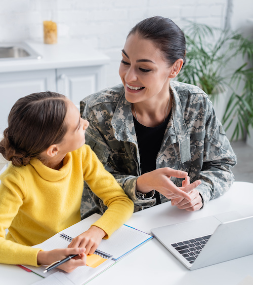 Cheerful woman in military uniform pointing at laptop near daughter and notebooks and home